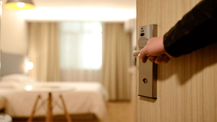 Hand opening a hotel room door with view of bed and furniture inside